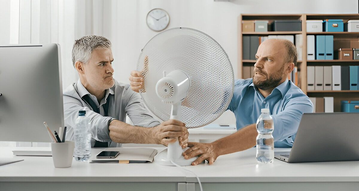 Two men struggling over a desk fan in an office setting, emphasizing the need for effective cooling solutions during hot weather, relevant to AC repair and maintenance services in Bucks County.